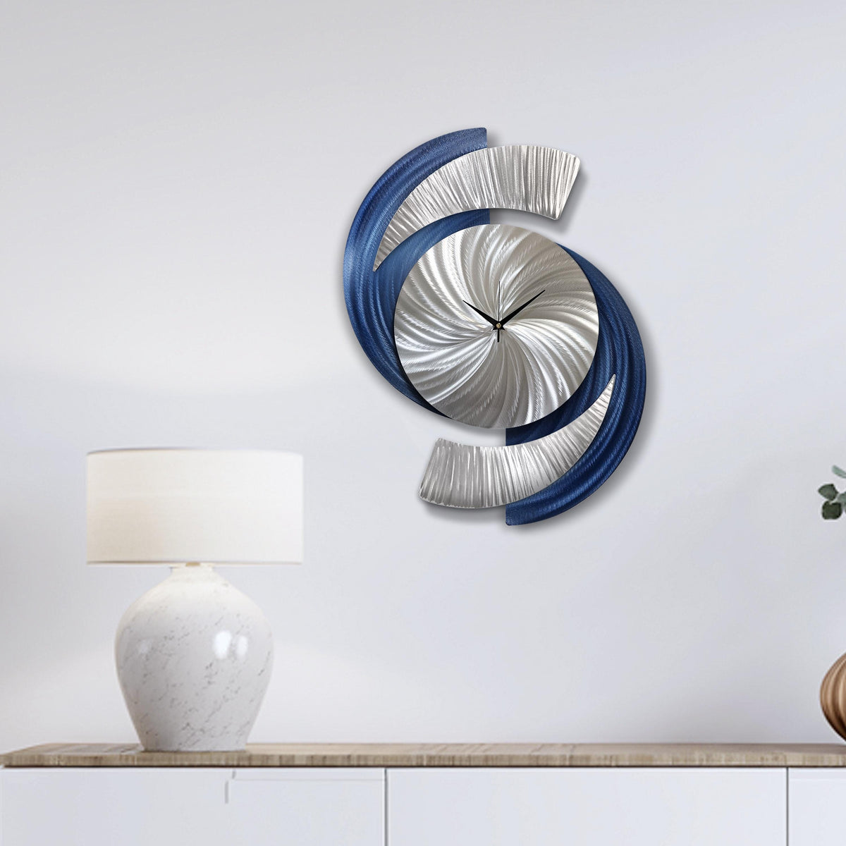 May include: Contemporary wall clock featuring a silver, blue, and grey abstract design. The clock face is silver with black hands. The clock is mounted on a white wall.