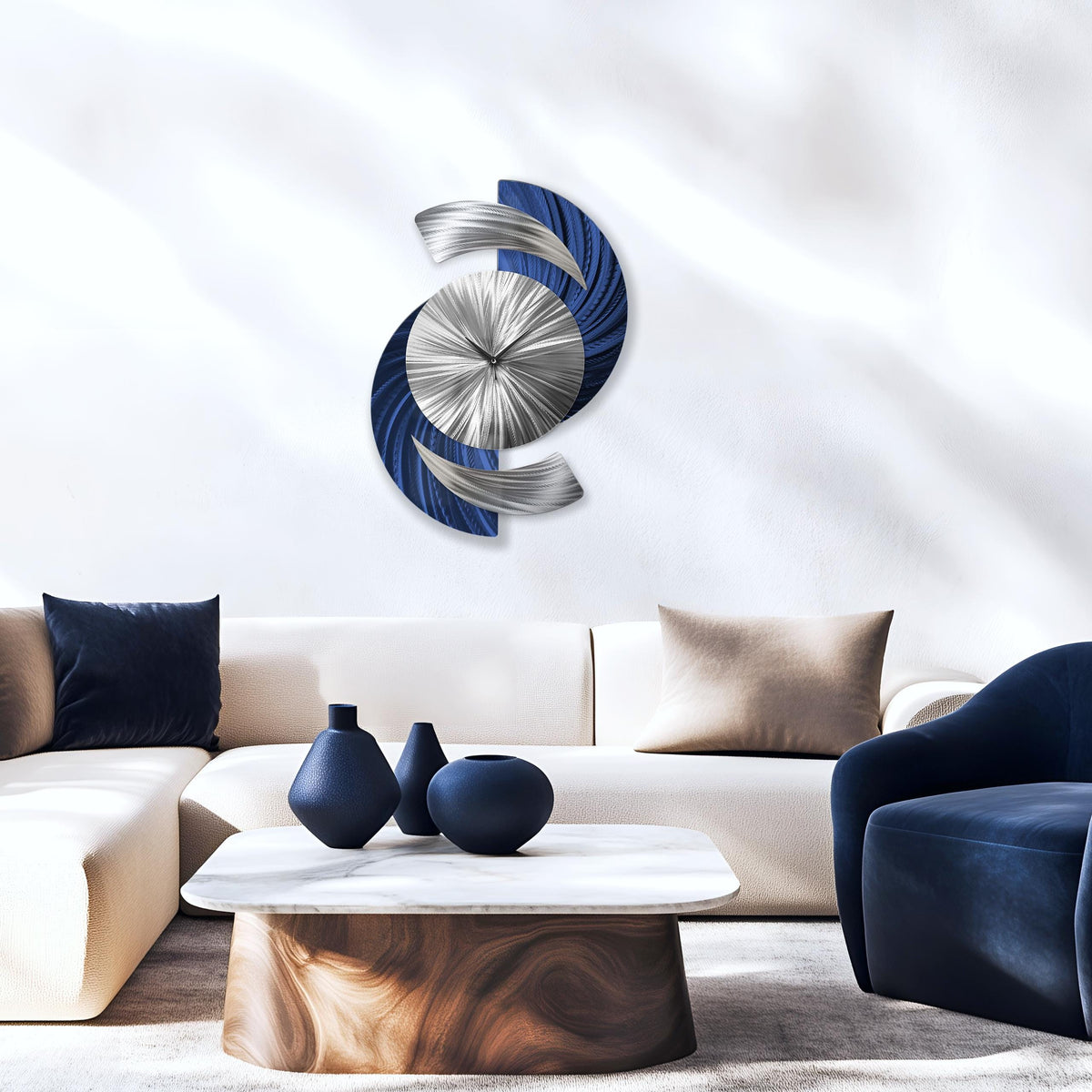 May include: A contemporary wall clock with a distinctive design. The clock has a silver central dial with radiating lines, framed by curved blue and silver metal elements. The clock is mounted on a white wall, above a white sofa and a marble coffee table.