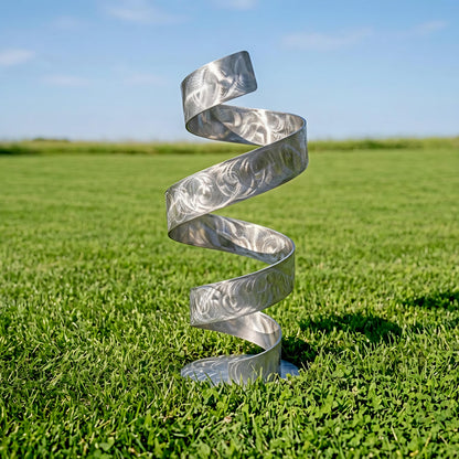 A silver spiral sculpture stands in a grassy field, its intricate design and metallic sheen contrasting with the natural surroundings.