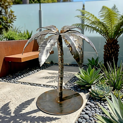 A metal sculpture of a palm tree stands in a garden, surrounded by various plants and rocks.