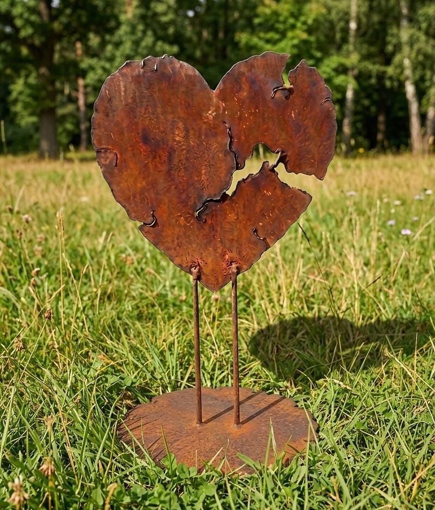 A rusted metal heart-shaped sculpture stands in a grassy field, with a tree in the background.