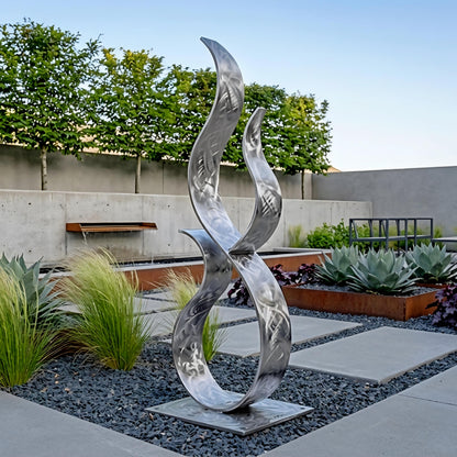 a modern garden sculpture with a unique, abstract design. The sculpture is made of metal and is situated in a garden setting with various plants and trees in the background.