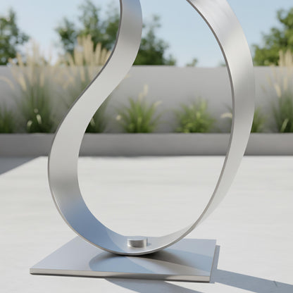 A modern, curved metal sculpture stands on a square base, with a blurred background of greenery and a clear blue sky.