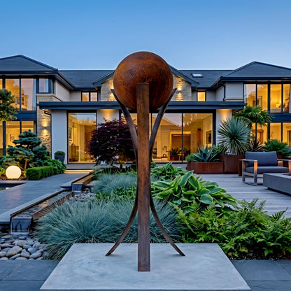 a large, modern house with a well-manicured garden in the foreground. The garden is adorned with various plants and a large, rustic sculpture that resembles a globe or ball. The house has a spacious deck and a pool, creating a serene and inviting atmosphere.