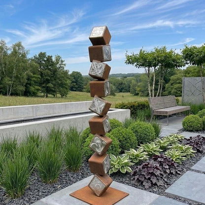 A tall, abstract sculpture composed of stacked cubes in various shades of brown and white, situated in a garden setting with lush greenery and a wooden bench.