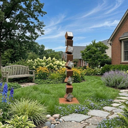 A tall, abstract sculpture made of stacked cubes stands in a lush, colorful garden, surrounded by various plants and flowers.