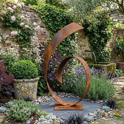 A large, rust-colored metal sculpture stands in a lush garden, surrounded by a variety of plants and flowers.