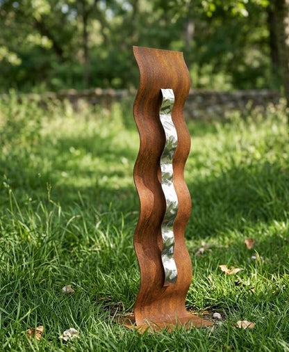 A tall, wavy metal sculpture stands in a grassy area, with a blurred background of trees.