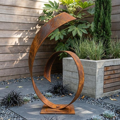 a large, rusted metal sculpture with a curved, metallic design, situated in a garden setting with plants and a wooden fence.