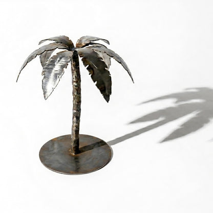 A metal sculpture of a palm tree stands on a round base, casting a shadow on the white background.