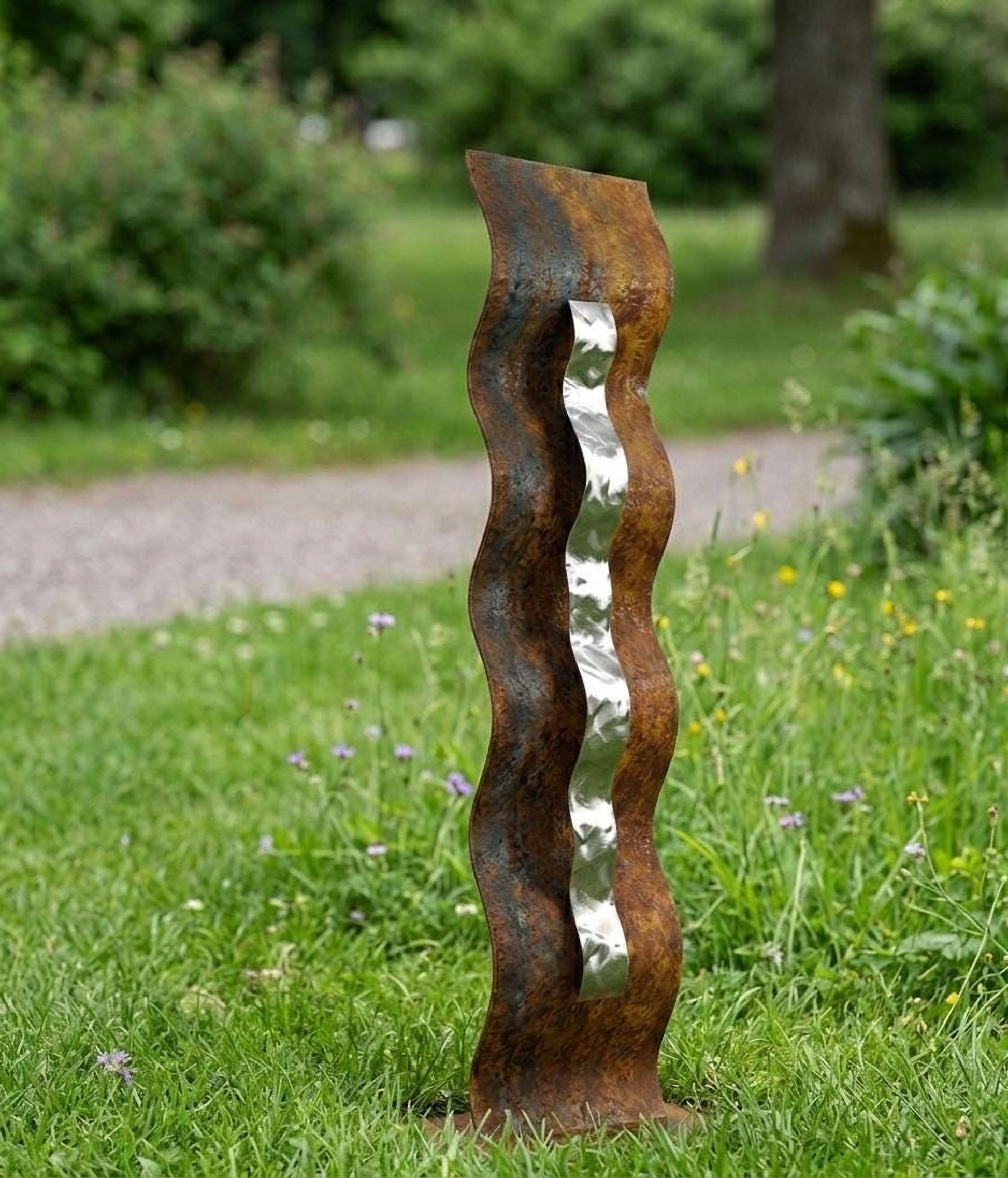A tall, rusted metal sculpture with a curved, metallic arm stands in a grassy area with trees and flowers in the background.