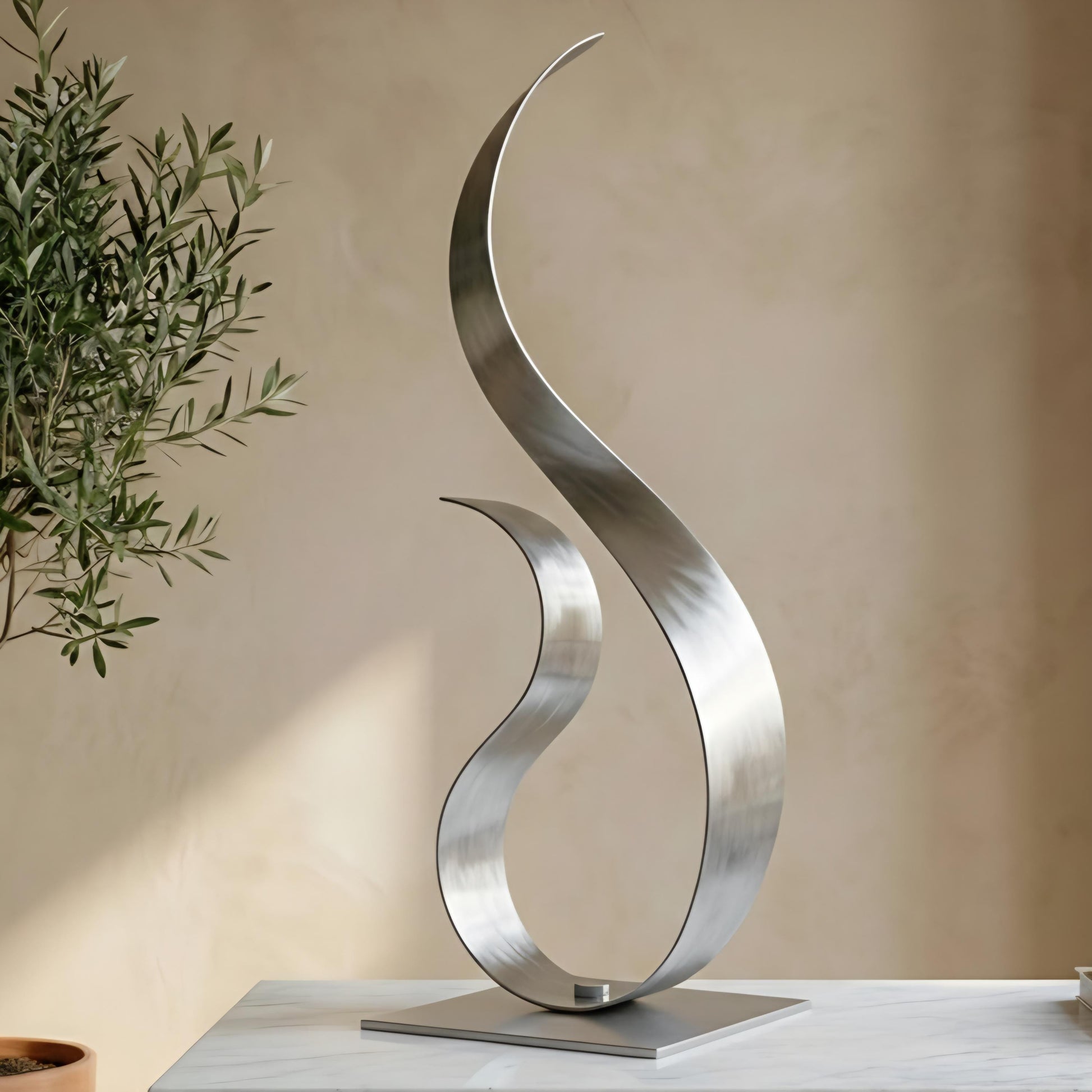 a modern, abstract sculpture made of metal, with a curved, metallic design that resembles a flame or a wave. The sculpture is placed on a white surface, and there is a potted plant visible in the background.