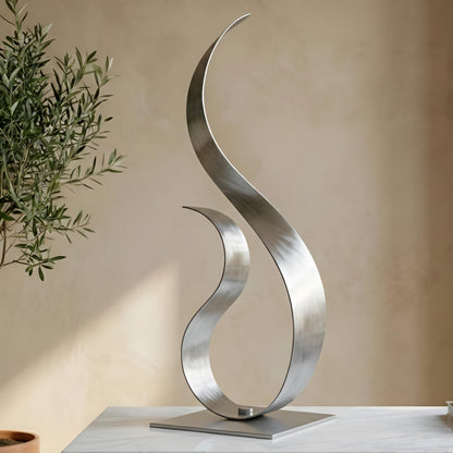 a modern, abstract sculpture made of metal, with a curved, metallic design that resembles a flame or a wave. The sculpture is placed on a white surface, and there is a potted plant visible in the background.