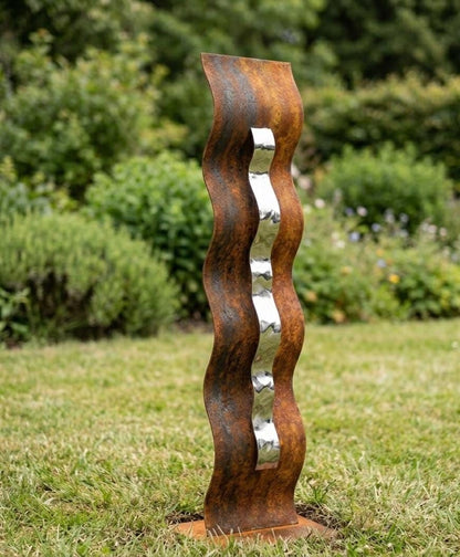 A tall, rust-colored metal sculpture with a curved, wavy design stands in a grassy area surrounded by greenery.