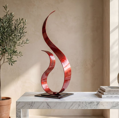 a modern red sculpture with a curved, metallic design, placed on a marble table. The sculpture is positioned next to a potted plant and a stack of books, creating a visually appealing and balanced composition.