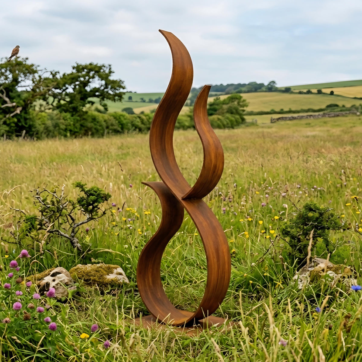 A large, abstract sculpture made of curved, brown metal stands in a grassy field with wildflowers and trees in the background.
