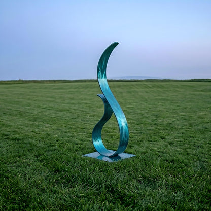 A blue metal sculpture resembling a flame stands in a grassy field, with a clear blue sky in the background.
