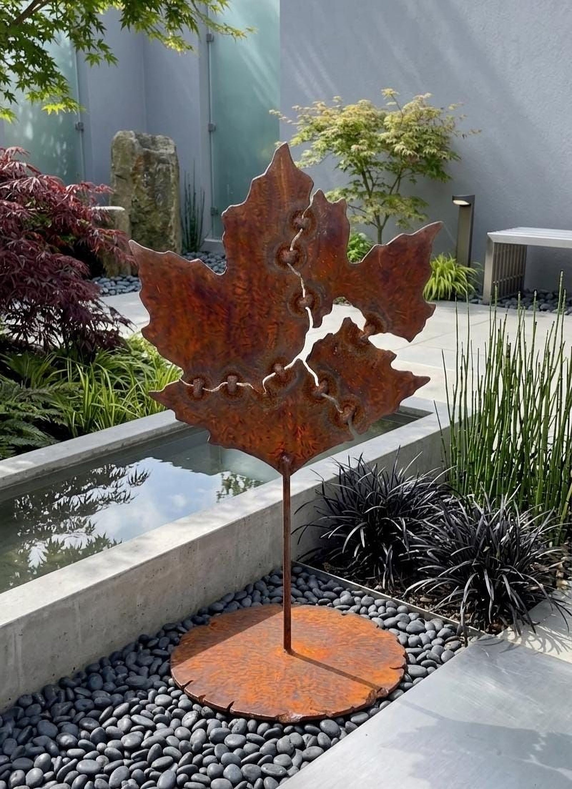 a rust-colored metal sculpture of a leaf, which is placed on a circular base and surrounded by rocks and plants.