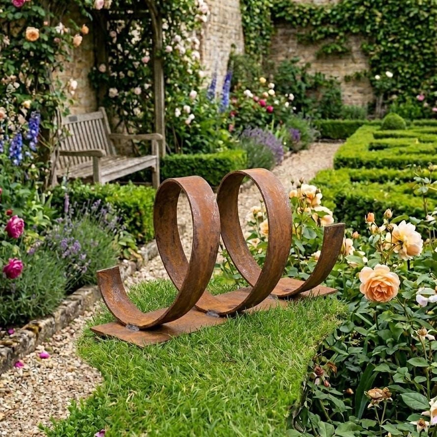 A rust-colored metal sculpture resembling a heart shape stands in a lush, well-maintained garden, surrounded by vibrant flowers and greenery.