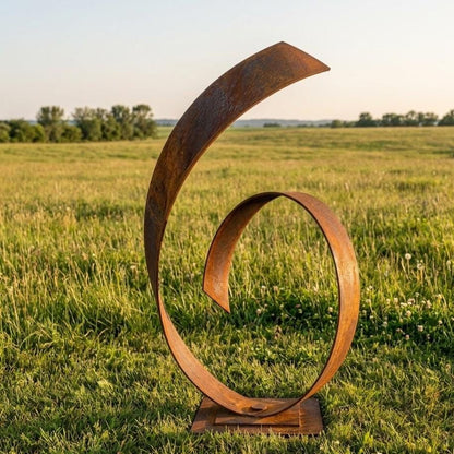A large, abstract metal sculpture resembling a twisted, curving shape stands in a grassy field, with a clear blue sky in the background.