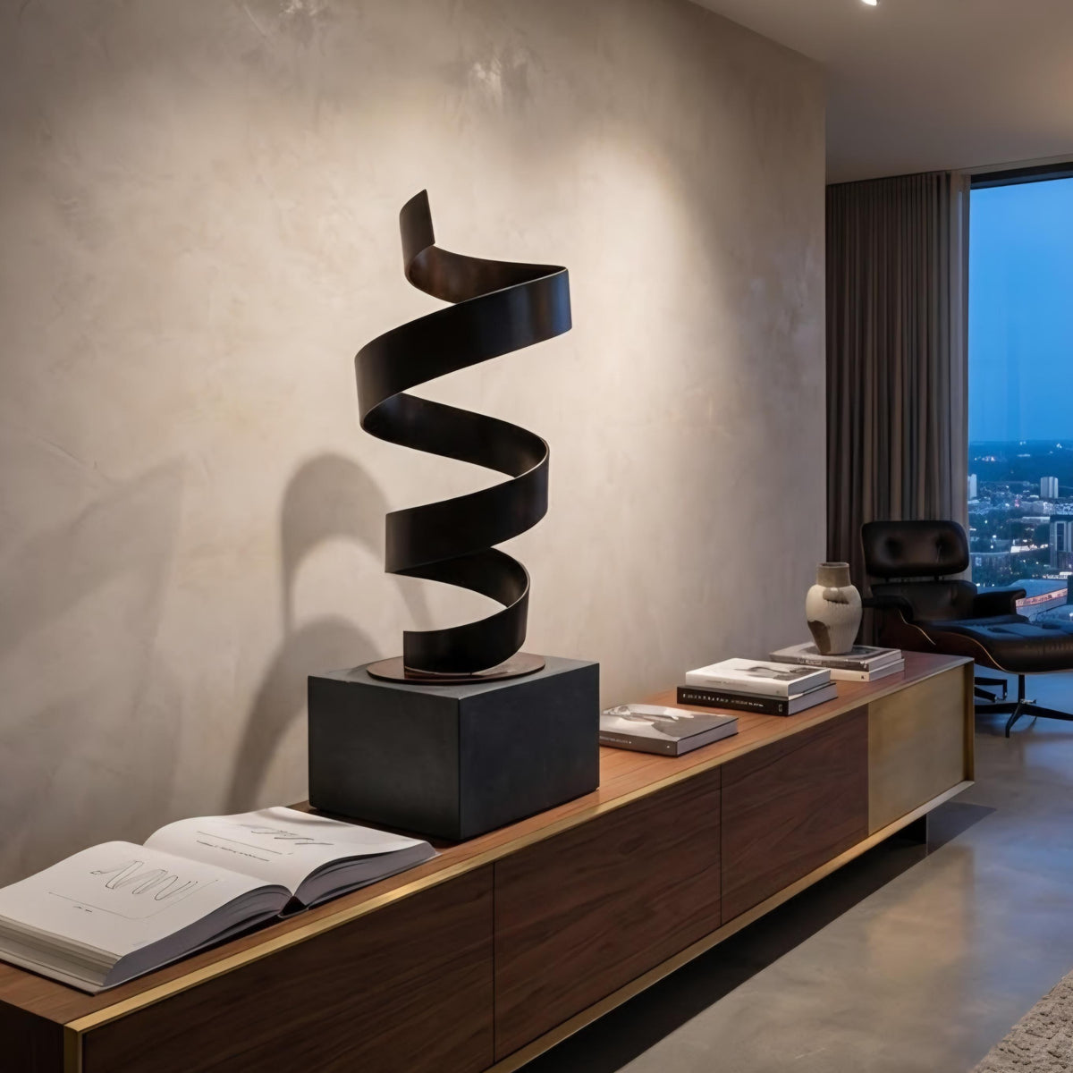 a modern living room with a wooden console table and a black spiral sculpture on top. The table also holds several books and a vase. The room has a large window overlooking a cityscape, and a leather chair is visible in the background.