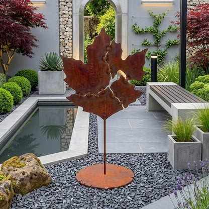 a garden with a large metal sculpture of a leaf in the center, surrounded by various plants and a small pond.
