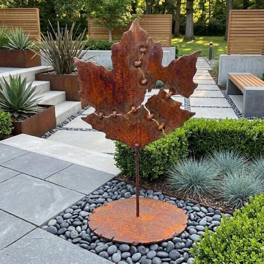 a large, rust-colored metal sculpture of a leaf, which appears to be a garden decoration. The sculpture is situated in a garden setting with various plants and rocks surrounding it.