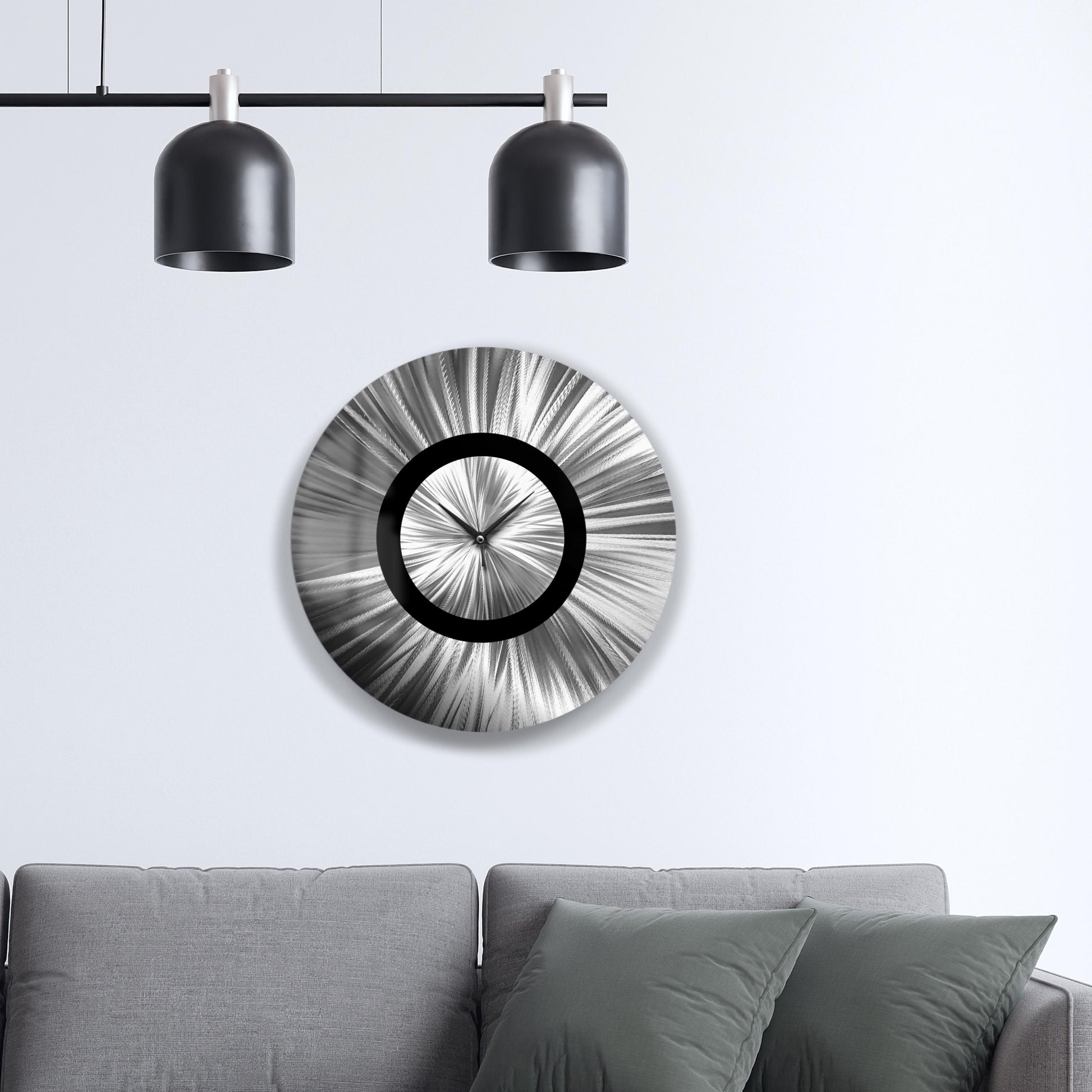 May include: A contemporary, circular wall clock featuring a silver radial pattern and a black central ring. The clock hands are black. The clock is mounted on a white wall above a grey sofa.