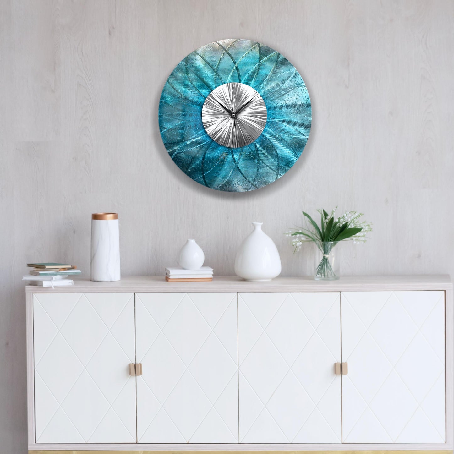 May include: A round, teal metal wall clock with a silver centre and hands. The clock has a textured, floral-like design. The clock is mounted on a light grey wall above a white cabinet with decorative doors.