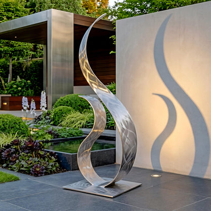 a modern, metallic sculpture with a unique, curved design, situated in a garden setting with a pond and lush greenery in the background.