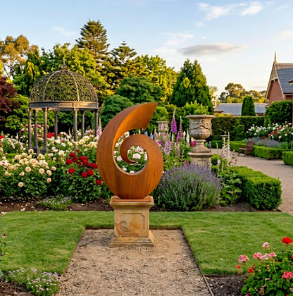 A large, ornate metal sculpture stands in a well-manicured garden, surrounded by colorful flowers and lush greenery.