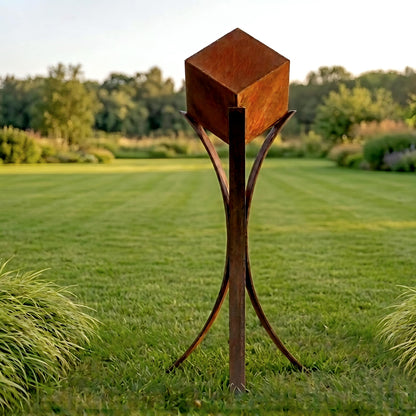 A large, rust-colored metal sculpture stands in a grassy field, surrounded by trees and shrubs.