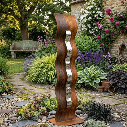 A tall, rust-colored metal sculpture with a curved, wavy design stands in a lush garden surrounded by various plants, flowers, and a stone path.