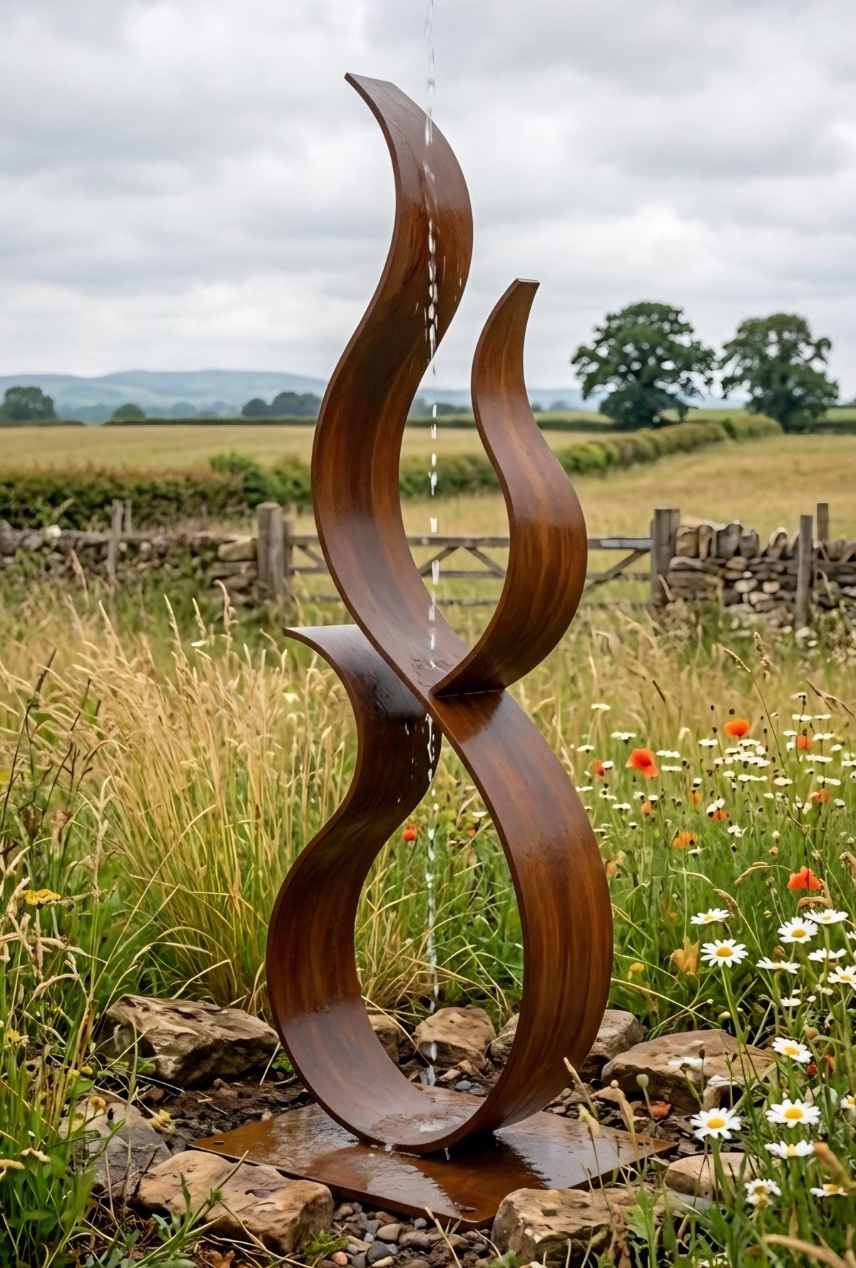 A large, abstract sculpture made of curved, dark brown metal stands in a field with wildflowers and a stone wall in the background.