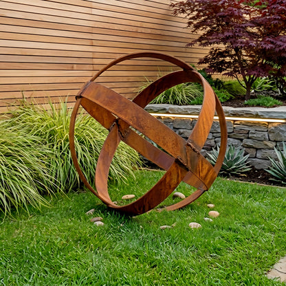 A large, rusted metal sculpture resembling a sphere or planet stands in a well-maintained garden, surrounded by lush greenery and a stone wall.
