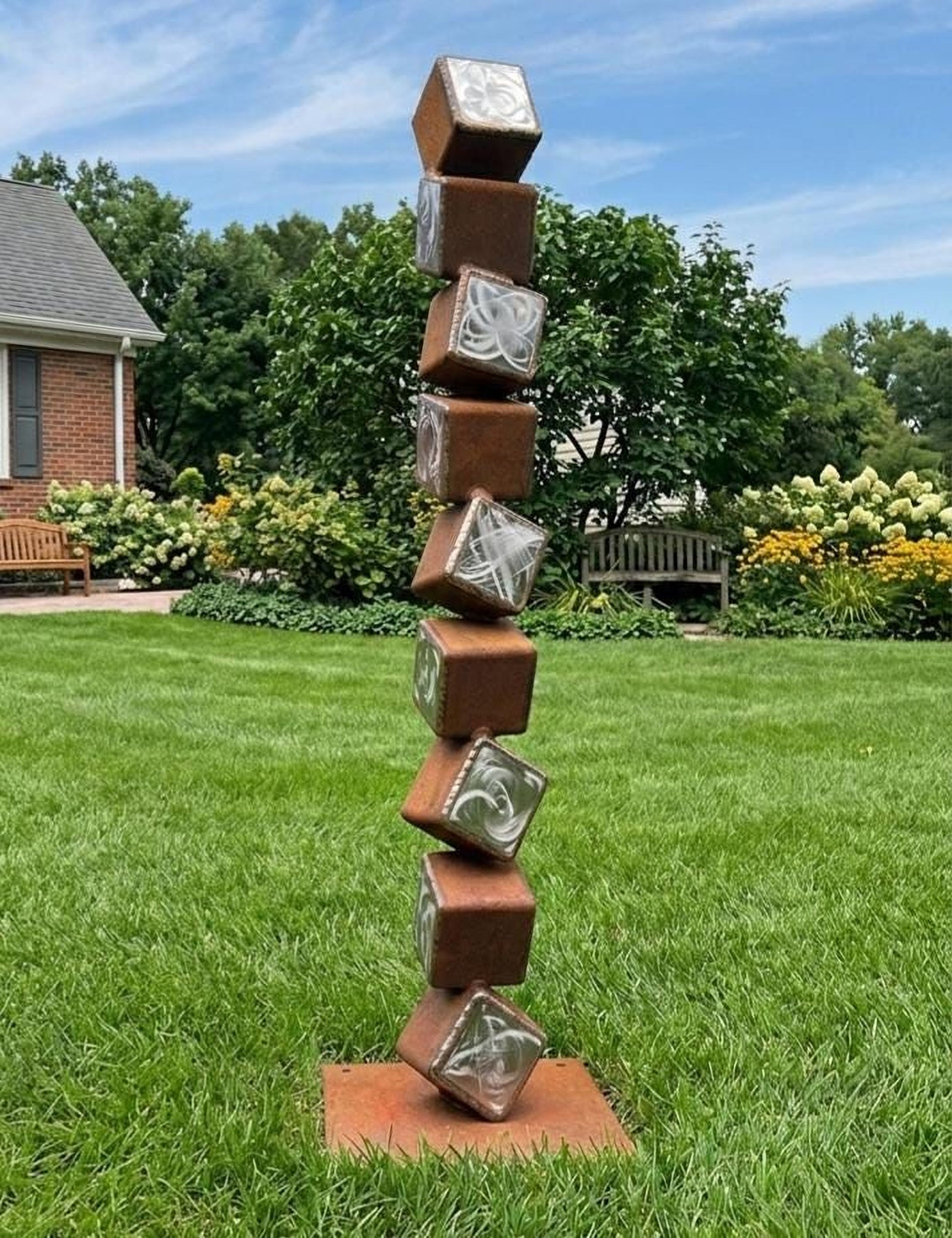 A tall, rust-colored metal sculpture composed of stacked cubes stands in a grassy yard, with a brick building and trees in the background.