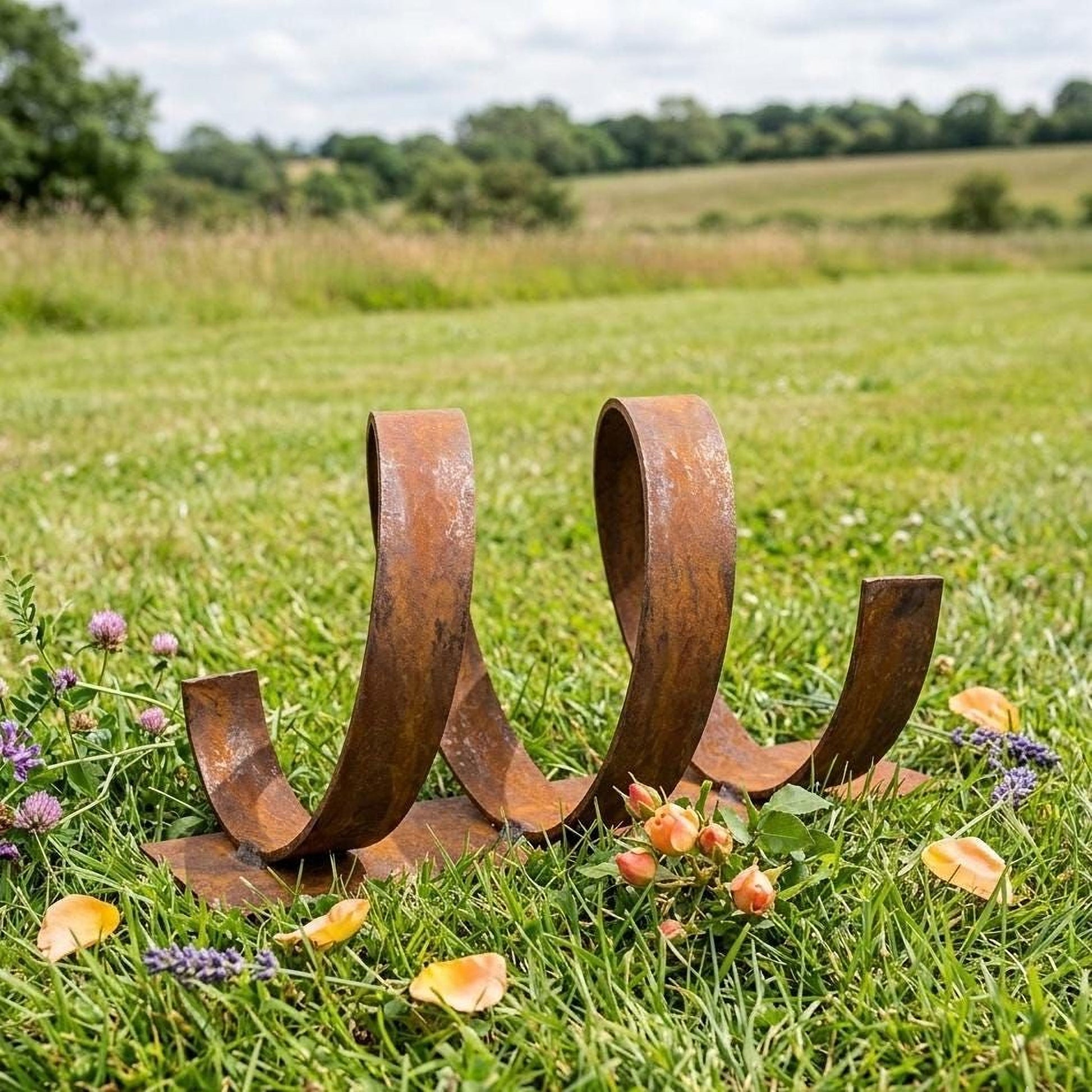 A rusted metal sculpture resembling a coiled spring rests in a grassy field, surrounded by purple flowers and yellow petals.