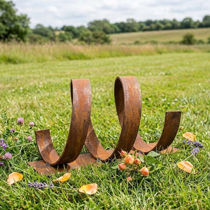 A rusted metal sculpture resembling a coiled spring rests in a grassy field, surrounded by purple flowers and yellow petals.