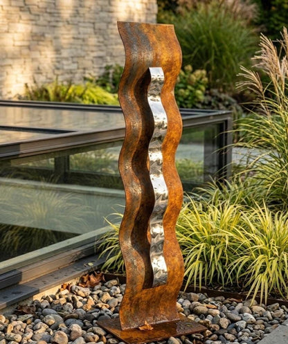a tall, rust-colored metal sculpture with a curved, metallic arm extending upwards, situated in a garden setting with greenery and a glass structure in the background.