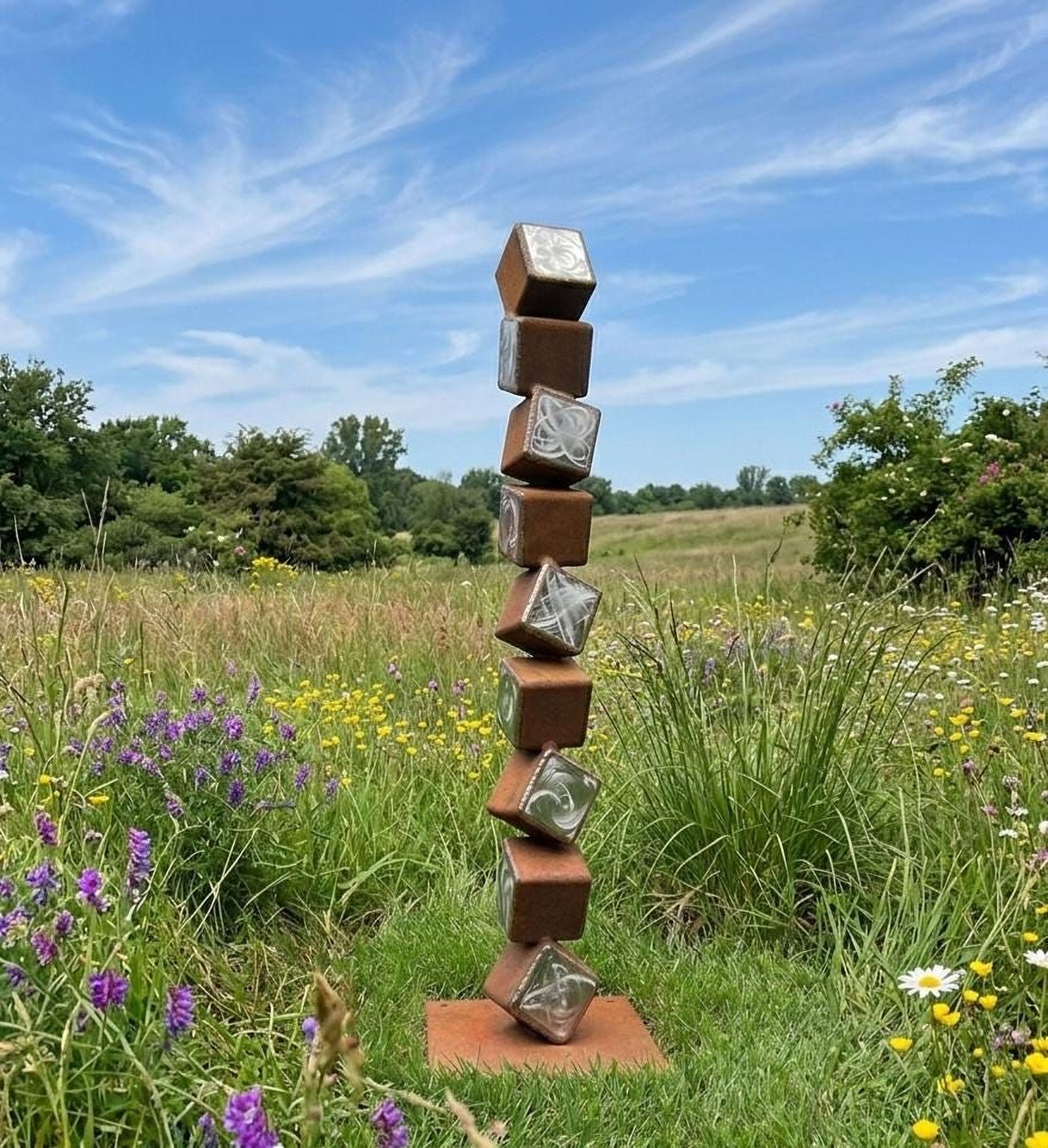 A tall, rust-colored sculpture composed of stacked cubes stands in a field of wildflowers, with a blue sky and trees in the background.