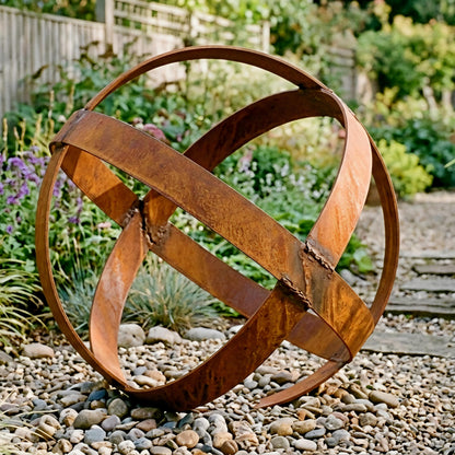 A large, rusted metal sculpture resembling a sphere or ball is situated in a garden setting, surrounded by rocks and greenery.