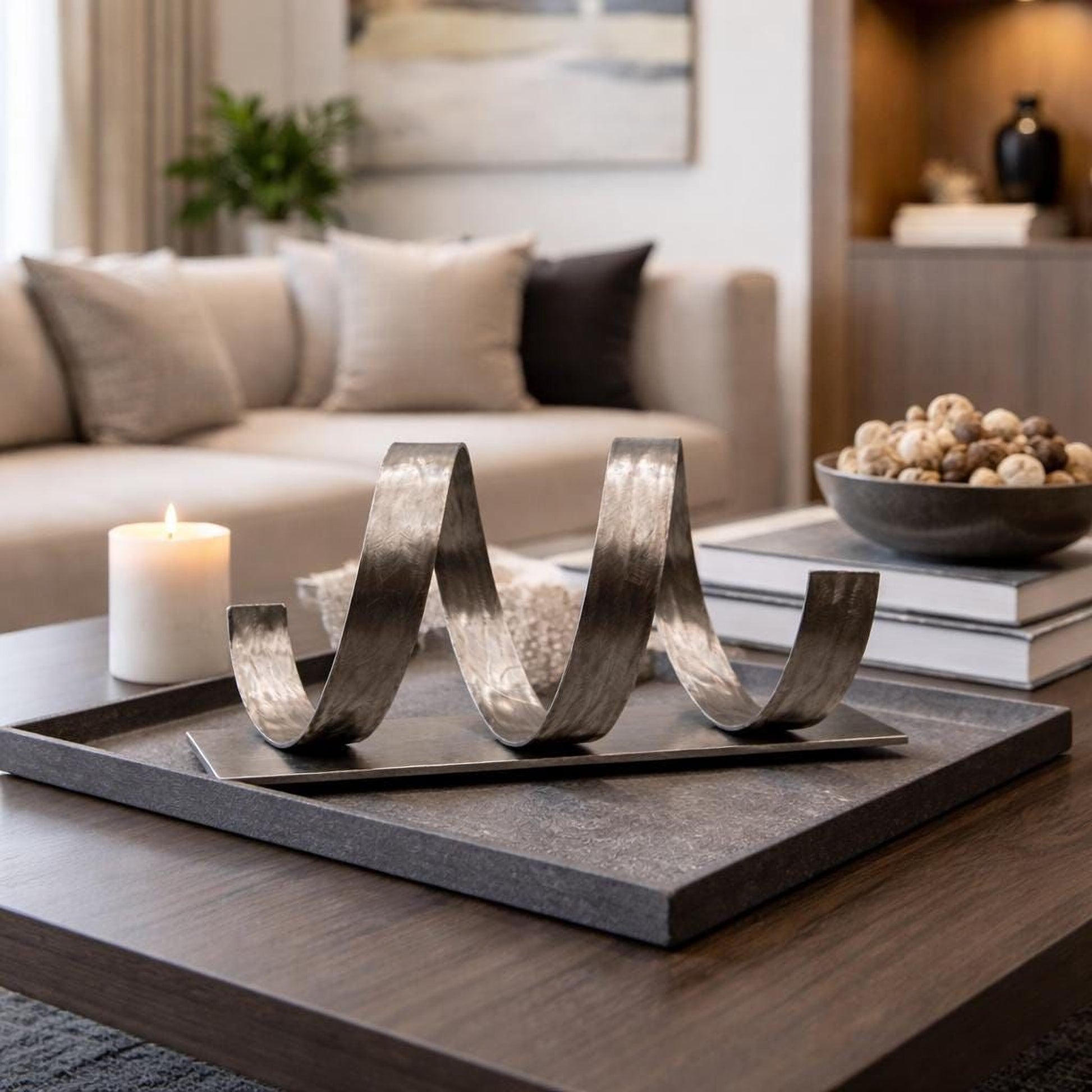 a modern living room with a wooden coffee table in the foreground, adorned with a decorative metal sculpture and a lit candle. The room is furnished with a couch, a bookshelf, and a plant, creating a cozy and inviting atmosphere.