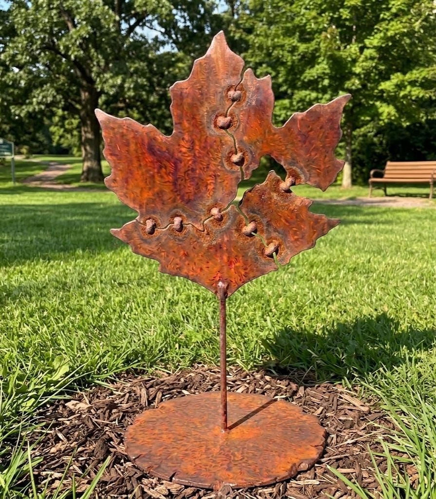 A rust-colored metal sculpture of a leaf stands on a bed of mulch in a park, surrounded by lush greenery.