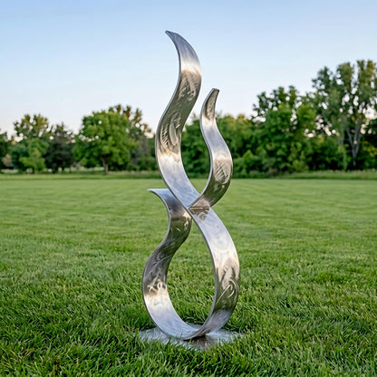 A large, abstract metal sculpture resembling a flame or fire stands in a grassy field, with trees visible in the background.