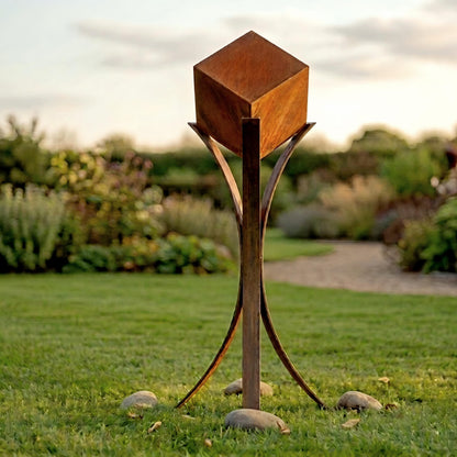 A large, square metal sculpture stands in a grassy area, surrounded by rocks.