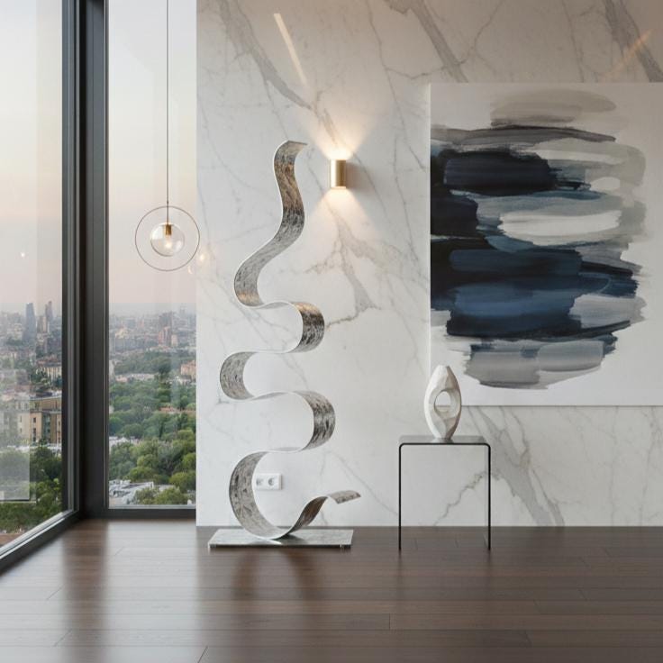 May include: A contemporary interior with a large, abstract metal sculpture in a ribbon design. The silver sculpture stands on a rectangular base. A painting with blue and grey brushstrokes hangs on the marble wall.
