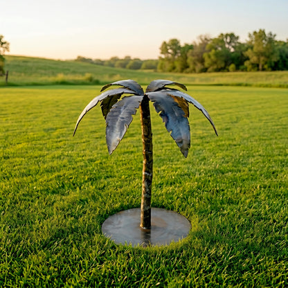 A metal sculpture of a palm tree stands in a grassy field, with a clear blue sky and trees in the background.
