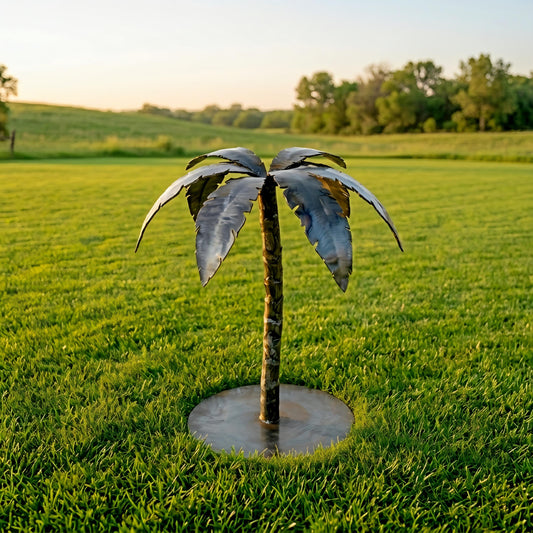 A metal sculpture of a palm tree stands in a grassy field, with a clear blue sky and trees in the background.