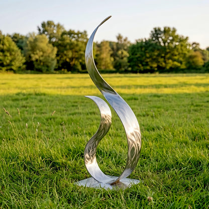 A large, metallic sculpture resembling a flame stands in a grassy field, with trees visible in the background.
